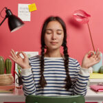 relaxed-asian-woman-meditates-workplace-sits-zen-pose-against-desktop-with-flowers-desk-lamp-notepads-wears-striped-casual-jumper-tries-relax-after-work-isolated-pink-background