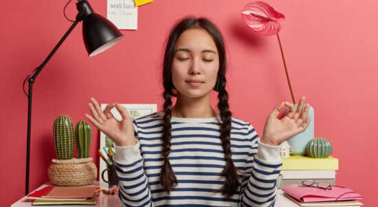 relaxed-asian-woman-meditates-workplace-sits-zen-pose-against-desktop-with-flowers-desk-lamp-notepads-wears-striped-casual-jumper-tries-relax-after-work-isolated-pink-background